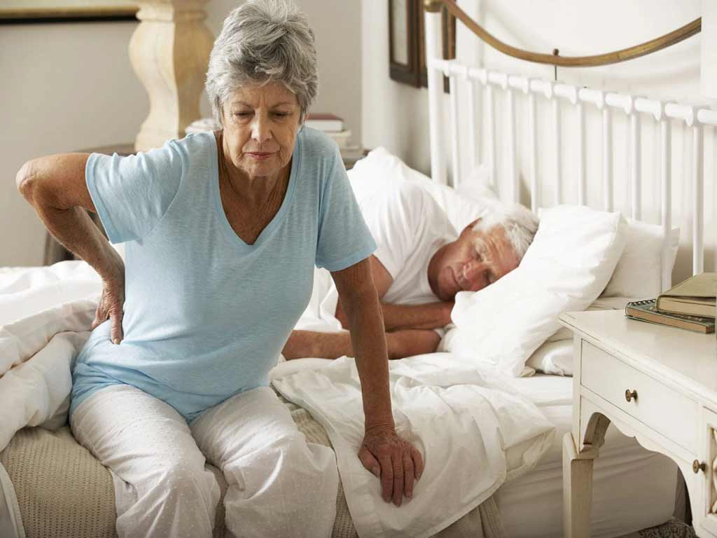 An elderly woman sitting on her bed touching her pained lower back