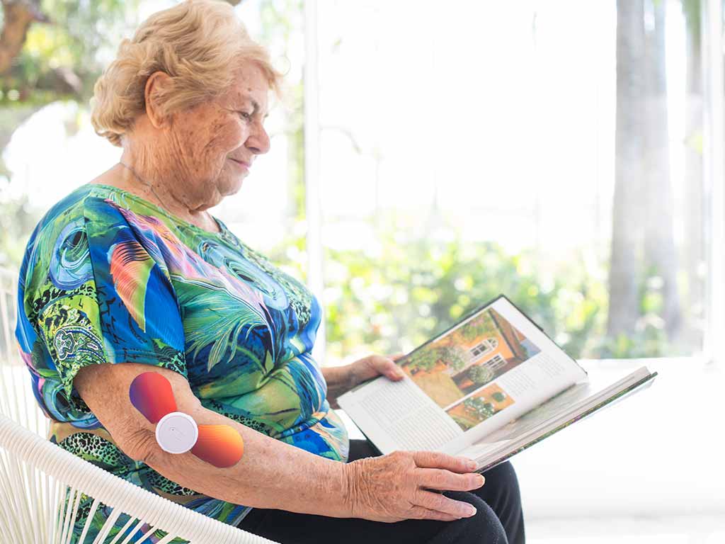 An elderly woman using a wireless TENS unit on her arm while reading.