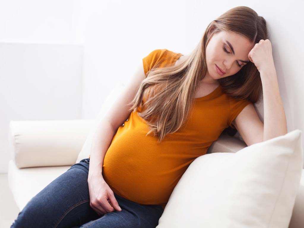 A sad pregnant woman sitting on her couch.