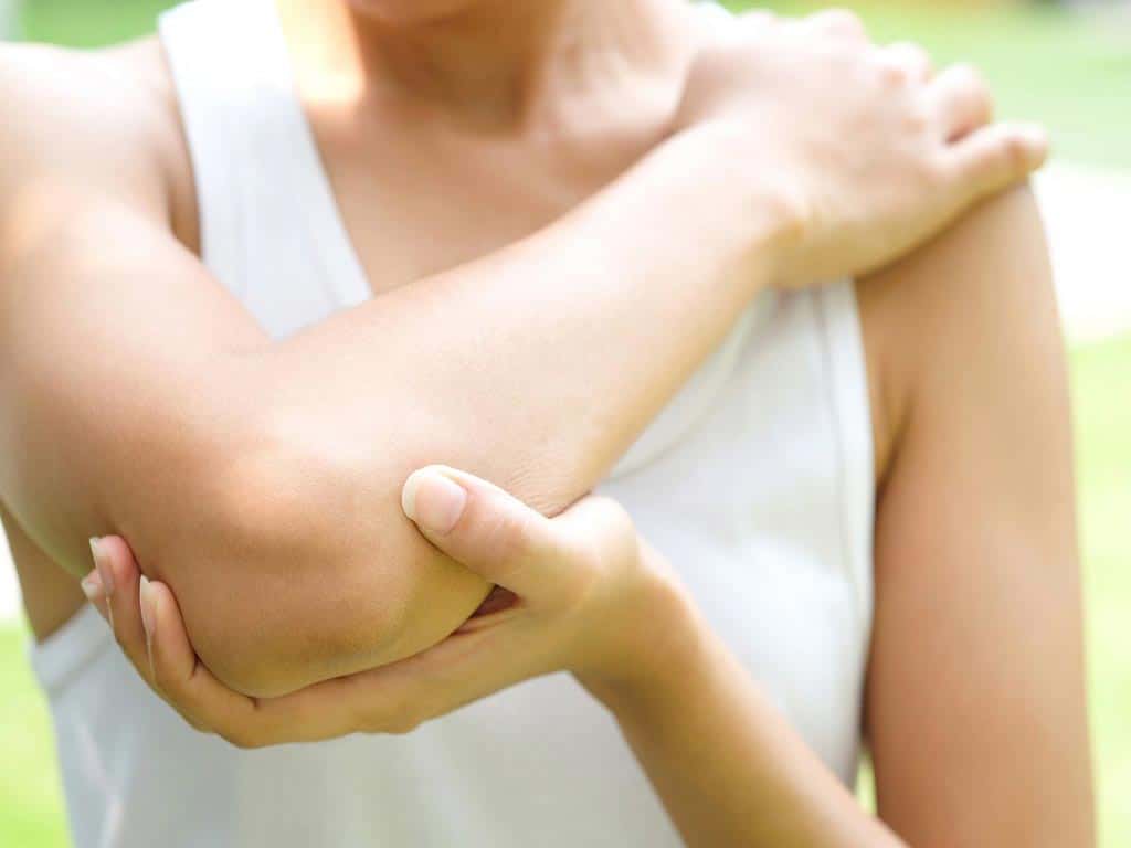 A woman wearing a white tank top is holiding her elbow