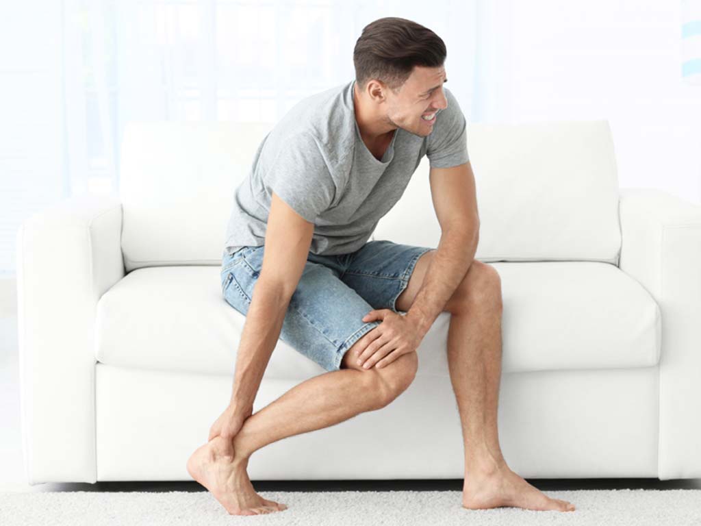 A man sitting on a couch while touching aching knee and foot