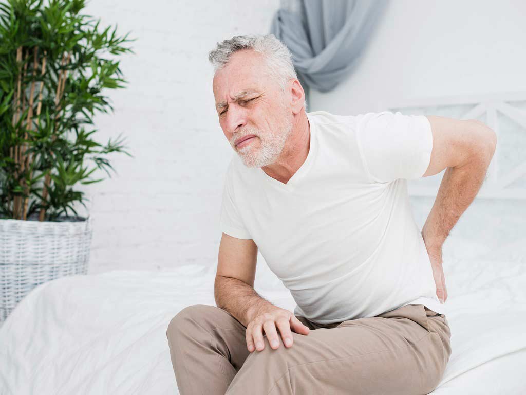 An elderly man sitting on a couch touching his painful lower back