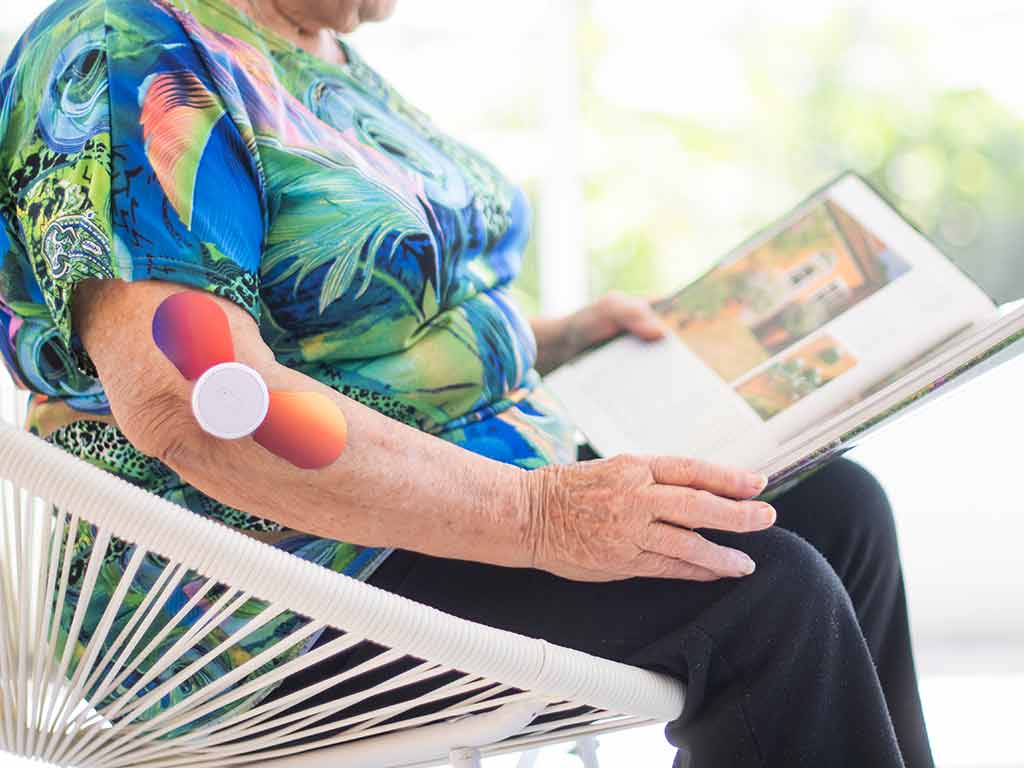 An elderly woman sitting on a chair while using a TENS machine of her arm and reading a book
